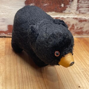 Peut inclure: Un ours en peluche recouvert d'un tissu noir texturé avec un museau beige lisse et de petits yeux ronds marron. L'ours est représenté en position debout avec des oreilles visibles et un corps arrondi, présentant une texture extérieure douce.