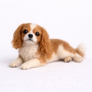 Op de afbeelding: Handgemaakte beeldje van een Cavalier King Charles Spaniel hond. De hond ligt, met een wit en bruin vacht, en grote, donkere ogen. Het beeldje heeft een pluizige staart en staat tegen een witte achtergrond.