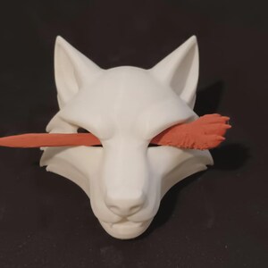 May include: A white wolf mask featuring a smooth, matte finish with pointed ears and a sharp snout. A long, slender, textured orange object is positioned horizontally across the eye openings, extending outward from both sides of the mask.