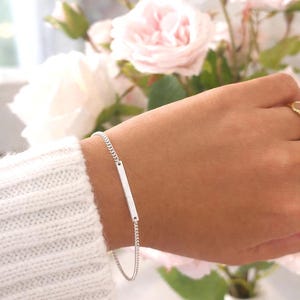 May include: A silver bar bracelet with a fine chain. The bracelet is displayed on a wrist, with a white sweater sleeve visible. Soft pink roses are in the background.