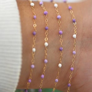 May include: Close-up of gold chain bracelets featuring small, round, purple and white beads. The bracelets are displayed on a wrist, highlighting the jewellery's design and colour combination. The beads are evenly spaced along the chains.