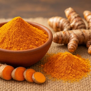 Puede incluir: Un cuenco de terracota lleno de polvo de cúrcuma de color naranja brillante, con un montón más pequeño de polvo al lado. También se ven raíces de cúrcuma en rodajas y raíces de cúrcuma enteras, todo sobre una superficie marrón texturizada.