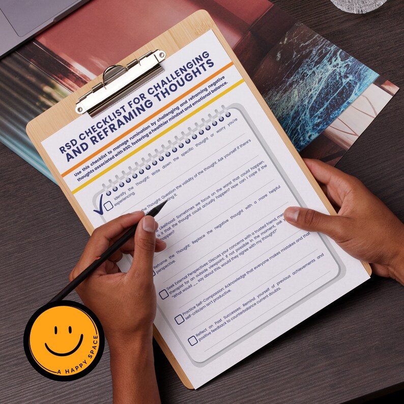 May include: A wooden clipboard with a checklist titled "RSD Checklist for Challenging and Reframing Thoughts." The checklist is being filled out with a black pen. A round sticker with a smiling face and the words "A Happy Space" is in the lower left corner.
