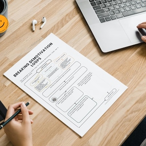 Puede incluir: Una hoja de trabajo titulada "Breaking Demotivation Loops" est&aacute; sobre un escritorio de madera, junto a un port&aacute;til y un cuaderno. Una mano sostiene un bol&iacute;grafo y otra usa el panel t&aacute;ctil del port&aacute;til.