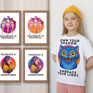Puede incluir: Una persona joven lleva una camiseta blanca con un gato azul y el texto "OWN YOUR SHADOW, EMBRACE YOUR LIGHT." Cuatro impresiones enmarcadas con ilustraciones y texto: "DIFFERENT IS POWERFUL" y "FEARLESS & UNDEFINED."