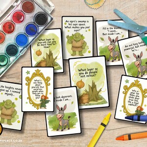 May include: A collection of illustrated cards with inspirational quotes and cartoon characters, including a green ogre and a donkey. The cards are arranged with a watercolor paint set, scissors, and crayons on a wooden surface.
