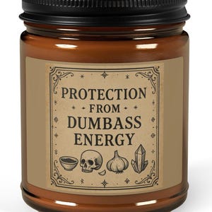 May include: A brown glass jar with a black lid. The label reads "PROTECTION FROM DUMBASS ENERGY" with illustrations of a skull, garlic, crystal, and bowl. The jar is filled with a light brown substance.