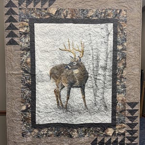 May include: A brown and white quilt with a camouflage border and a white deer with antlers in the center. The deer is standing in a snowy forest setting.