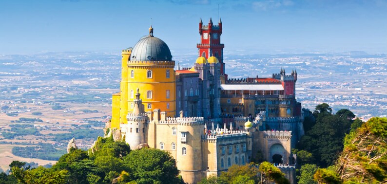 May include: A yellow and red castle with a large dome on top, set against a blue sky and a distant view of a city.