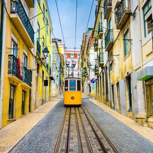 May include: A yellow vintage streetcar travels up a steep, cobblestone street lined with colorful buildings in Lisbon, Portugal.