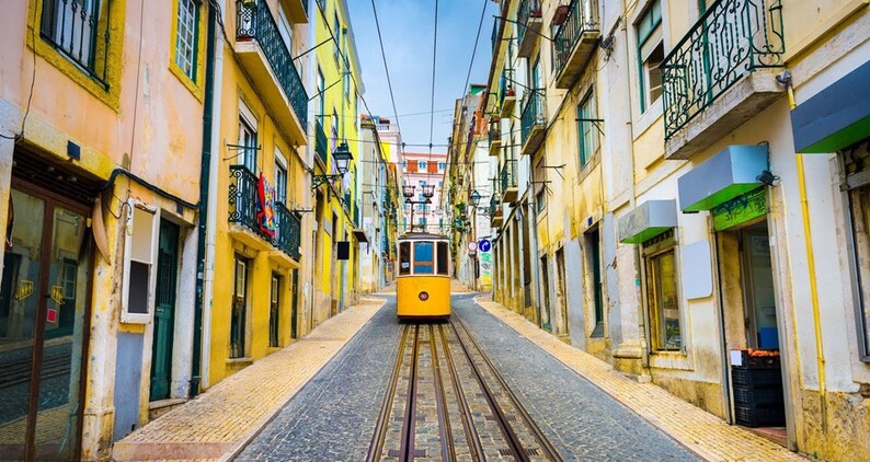May include: A yellow vintage streetcar travels up a steep, cobblestone street lined with colorful buildings in Lisbon, Portugal.