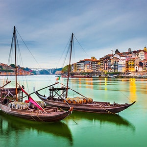 May include: Three traditional Portuguese boats called Rabelos, used for transporting port wine, are docked in the Douro River in Porto, Portugal. The boats are made of wood and have a distinctive shape with a high bow and stern. The boats are painted in a variety of colors, including red, white, and blue. The boats are docked in front of a cityscape with buildings on the waterfront.