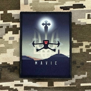 Pode incluir: Um remendo bordado preto e azul com um drone voando sobre um tridente ucraniano estilizado. O drone tem luzes vermelhas e o texto "MAVIC" está impresso abaixo do drone.