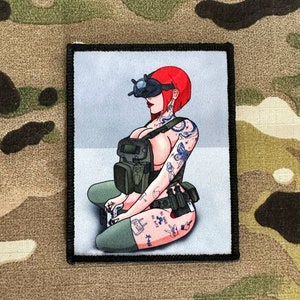 Könnte beinhalten: Ein schwarz-weißes Patch mit einer Comic-Illustration einer Frau, die ein Virtual-Reality-Headset und taktische Ausrüstung trägt. Sie hat rote Haare und Tattoos. Der Text "KILL DEAD ALL BUTTE" ist auf ihrem Bein sichtbar.