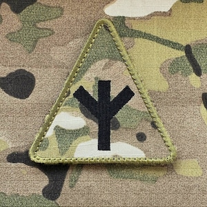 Pode incluir: Um tecido de camuflagem preto e verde com um remendo triangular verde. O remendo tem um símbolo preto no interior, que se assemelha a uma árvore estilizada.