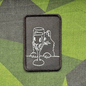 Puede incluir: Un parche bordado negro con un contorno blanco de un gato mirando por encima de un mostrador con una copa de champán.