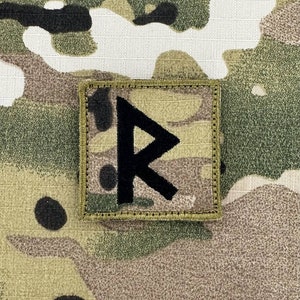 以下が含まれることがあります： 迷彩柄の背景に黒いルーン文字のシンボルが描かれた正方形のパッチ。 パッチはベージュの縁取りがあり、おそらく布製です。 迷彩柄は、緑、茶色、ベージュの色合いを含んでいます。