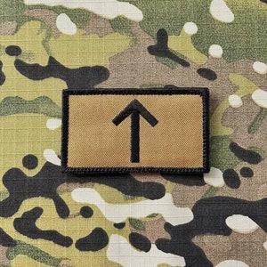 以下が含まれることがあります： 上向きに指し示す黒い矢印が描かれた、薄い茶色の布製のワッペン。ワッペンは黒い縁取りがされている。