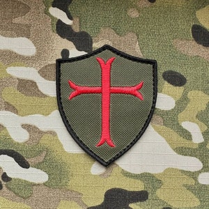 May include: A green fabric patch shaped like a shield with a red cross in the center. The patch has black stitching around the edges.