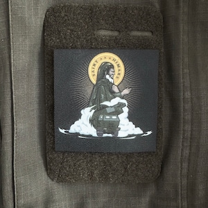 Könnte beinhalten: Ein schwarzer Patch mit einem weißen und goldenen Bild einer Person in einer Robe mit den Worten "Saint Himars" um das Bild herum.