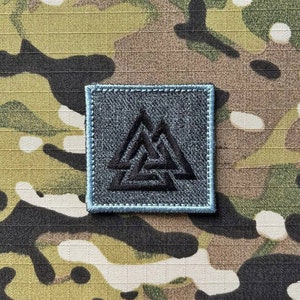 May include: A black and gray embroidered patch with a triangular symbol. The patch has a blue border and is sewn onto a camouflage fabric.
