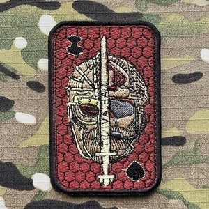 Może przedstawiać: Czerwona naszywka haftowana z czarnym piko w prawym dolnym rogu. Naszywka przedstawia stylizowany obraz hełmu z przechodzącym przez niego mieczem.