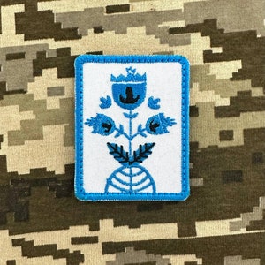 Op de afbeelding: Een witte geborduurde patch met een blauwe rand. De patch heeft een gestileerde blauwe bloem met een zwarte stengel en bladeren. De bloem heeft een kroon bovenop.