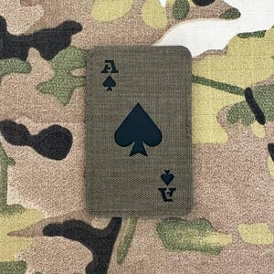 Könnte beinhalten: Ein rechteckiger Aufnäher mit dem Design des Pik-Ass. Der Aufnäher hat einen olivgrünen Hintergrund mit einem dunkelblauen Pik-Symbol und dem Buchstaben "A". Der Aufnäher ist vor einem Tarnmuster platziert.