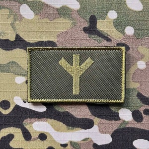 以下が含まれることがあります： 緑と茶色の迷彩柄の生地のパッチに、黄色の刺繍でシンボルが施されています。シンボルは、3つのポイントを持つスタイリッシュなY字型です。