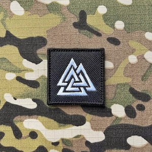 May include: A black patch with a white embroidered Valknut symbol. The patch is on a camouflage background.