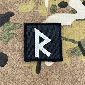 以下が含まれることがあります： 黒い布のパッチに白い刺繍のルーンシンボルが施されています。ルーンはスタイリッシュな文字「R」です。