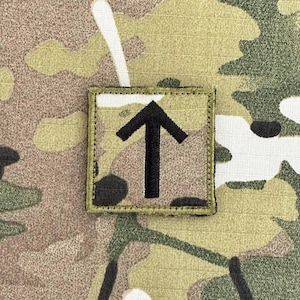 Può includere: Un cerotto di tessuto mimetico verde e beige con una freccia nera ricamata che punta verso l'alto.