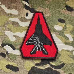 Pode incluir: Um remendo bordado vermelho e preto em forma de triângulo com uma silhueta preta de um caça dentro. O caça tem um contorno branco.