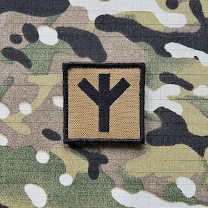 以下が含まれることがあります： ベージュの布地に黒い刺繍が施されたパッチ。シンボルは、上部に3つのポイントがあり、下に長い茎が伸びている、スタイリッシュなYの形をしています。