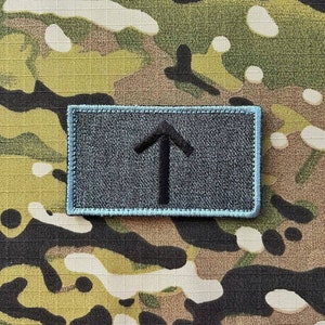 以下が含まれることがあります： 明るい青色の縁取りのある濃い灰色のパッチに、上向きに指し示す黒い矢印が描かれています