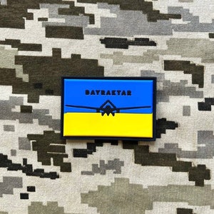 Könnte beinhalten: Ein blauer und gelber Patch mit dem Text "BAYRAKTAR" in schwarzen Buchstaben. Eine schwarze Silhouette einer Drohne ist mittig auf dem Patch platziert.