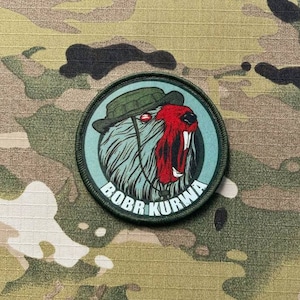 Könnte beinhalten: Ein runder Aufnäher mit einem Cartoon-Biber, der einen grünen Hut und einen roten Mund trägt. Der Biber ist vor einem türkisfarbenen Hintergrund mit den Worten "BOBR KURWA" in Weiß platziert. Der Aufnäher befindet sich auf einem Tarnhintergrund.