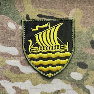 Könnte beinhalten: Ein schwarz-gelber, gestickter Aufnäher mit einem Wikingerschiff-Design. Der Aufnäher ist grün umrandet und vor einem Tarnmuster platziert.