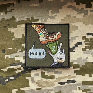 Peut inclure: Un patch brodé vert avec une image de dessin animé d'une botte piétinant une main avec la lettre "Z" dessus. La botte est marron et porte un drapeau jaune et bleu. La main est blanche et a une ongle rose. Une bulle de dialogue au-dessus de la botte dit "Put In!"