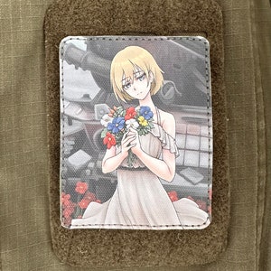 Può includere: Una toppa bianca con un'immagine di cartone animato di una giovane donna con i capelli biondi che tiene un mazzo di fiori. La donna indossa un vestito bianco con un collo a balze. Lo sfondo è un'immagine sfocata di una strada cittadina.