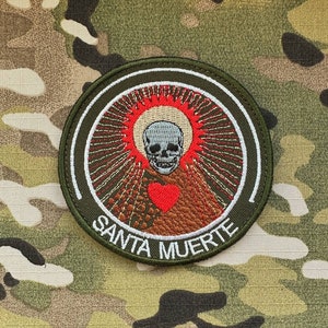 Könnte beinhalten: Runder, gestickter Aufnäher mit einem Totenkopf, einem roten Herzen und dem Schriftzug "SANTA MUERTE". Das Design umfasst einen Heiligenschein aus roten Strahlen und einen grünen Rand, vor einem Tarnmuster-Hintergrund.