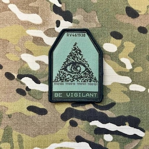 Op de afbeelding: Een zwart en groene patch met een witte achtergrond. De patch heeft een piramide met een oog erin en de tekst "RV461938" bovenaan. De tekst "BE VIGILANT" staat onderaan de patch. De piramide is gemaakt van een reeks zwarte en witte vierkanten.