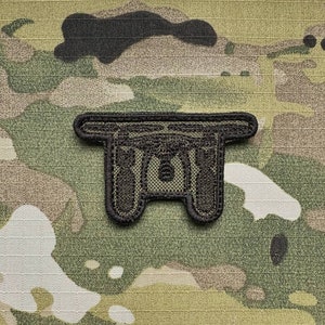 May include: A black and green embroidered patch with a stylized image of a military helmet. The patch is designed to be sewn onto clothing or gear.