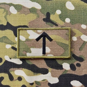 May include: A black arrow pointing upwards is embroidered on a tan fabric patch with a green border. The patch is on a camouflage background.