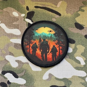 May include: A round black patch with a colorful image of three figures walking through a forest with a sunset in the background. The figures are silhouetted against the sunset and the forest is green and brown. There are two helicopters flying in the sky above the figures.