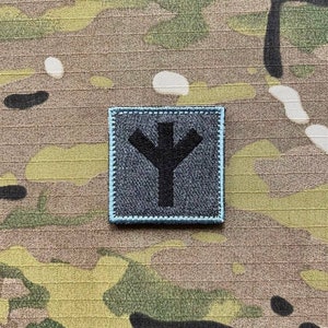 以下が含まれることがあります： グレーの背景にスタイリッシュな木のシンボルが描かれた白黒のパッチ。パッチは薄い青色の縁取りがあり、迷彩生地に縫い付けられています。