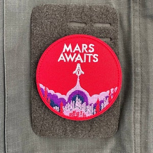 Può includere: Toppa circolare rossa con testo bianco "MARS AWAITS" e una silhouette bianca di un razzo. Lo sfondo è un paesaggio viola e rosa con alberi.