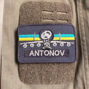 May include: A dark blue patch featuring a white silhouette of an aircraft with the text "ANTONOV" in white. The aircraft has yellow and blue stripes on its wings, similar to the Ukrainian flag.