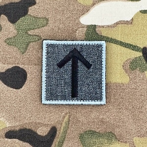 以下が含まれることがあります： 薄い青色の縁取りのある濃い灰色の正方形のパッチに、上向きに指し示す黒い矢印が刺繍されています。