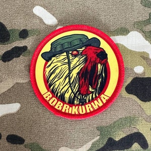 May include: A red and yellow embroidered patch with a cartoon image of a snarling animal wearing a green hat. The text "BOBR KURWA" is written in a yellow arc around the animal.